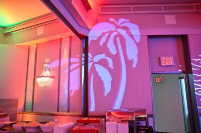 A brightly lit buffet area in NY with food trays and stacks of plates, featuring a palm tree light projection on the wall, two illuminated exit signs, and perfect for events with DJ services in Suffolk and Nassau County, NY.