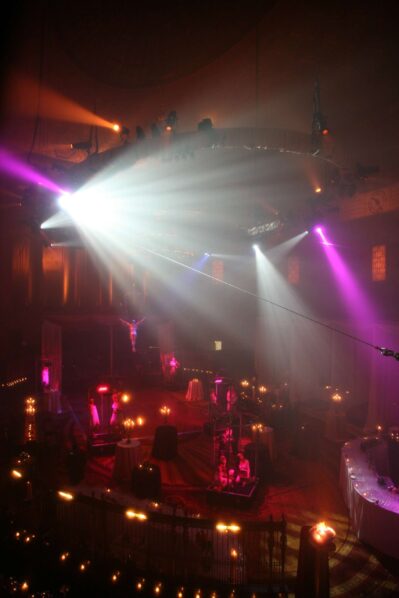 Dimly lit, elegant event hall in NY with dramatic spotlights in purple, pink, and white shining onto tables and a central stage. Round tables with candles and draped fabric create an atmospheric scene perfect for entertainment services Suffolk and Nassau County, NY.