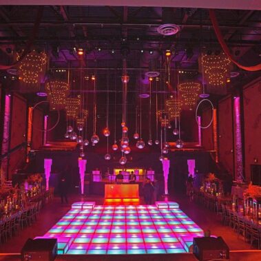 A large, dimly lit event space with a colorful illuminated dance floor, hanging disco balls, elegant table settings, and vibrant lighting—perfect for parties enhanced by top entertainment services Suffolk and Nassau County.