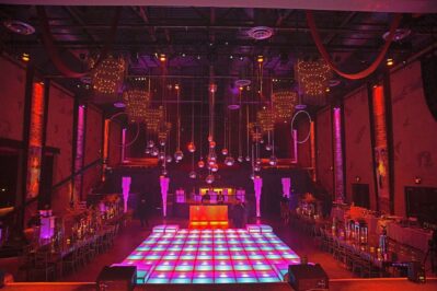 A large, dimly lit event space with a colorful illuminated dance floor, hanging disco balls, elegant table settings, and vibrant lighting—perfect for parties enhanced by top entertainment services Suffolk and Nassau County.