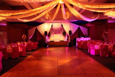A decorated banquet hall with draped white fabric and string lights on the ceiling, pink lighting, round tables with pink covers and bows, a stage with white chairs and florals—perfect for events featuring top entertainment services in Suffolk and Nassau County, NY.