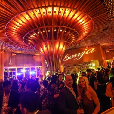 A large crowd socializes in an elegant, warmly lit NY bar with a striking ceiling design. The name “Sonja” glows above the bar as guests in formal attire enjoy the evening, complemented by top-notch DJ services Suffolk and Nassau County.