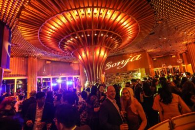 A large crowd socializes in an elegant, warmly lit NY bar with a striking ceiling design. The name “Sonja” glows above the bar as guests in formal attire enjoy the evening, complemented by top-notch DJ services Suffolk and Nassau County.