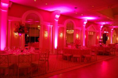 A banquet hall in NY decorated for an event, featuring round tables with chairs, elegant table settings, floral centerpieces, vibrant pink lighting, and top entertainment services Suffolk and Nassau County for a memorable celebration.