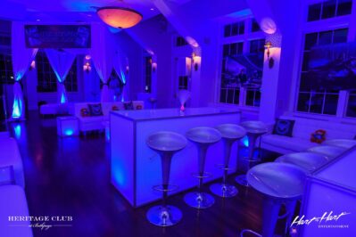 A modern NY lounge with glowing blue lighting, clear barstools, and white sofas. Plush toys and party decor add a festive touch. Large windows, wall art, and top DJ services in Suffolk and Nassau County complete the stylish setting.