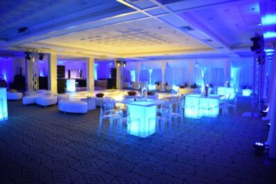 A spacious event hall with glowing blue lights, illuminated tables, transparent chairs, and white lounge seating—perfect for parties or receptions featuring top DJ services in Suffolk and Nassau County, NY—creates a modern and elegant atmosphere.