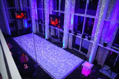 A modern indoor event space in NY with a white runway stage, purple and blue lighting, spotlights creating patterns on the floor, high ceilings, and large windows. Perfect for events needing DJ services Suffolk and Nassau County, NY. Tables and screens are also visible.