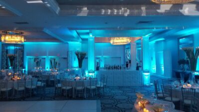 Elegant banquet hall with round tables, silver chairs, tall floral centerpieces, and glowing candles. Blue uplighting and a bar beneath a chandelier set the scene—perfect for events needing DJ services in Suffolk and Nassau County, NY.