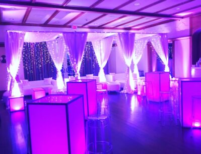 A lounge area is decorated with white drapes, fairy lights, and illuminated purple lighting. Clear bar stools and glowing tables sit on a wooden floor, creating a modern event space perfect for dj services Suffolk and Nassau County, NY.