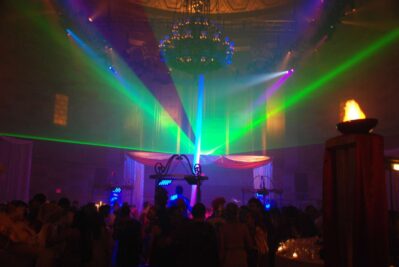 A crowded dance floor under a large chandelier, with colorful green and purple laser lights shining through fog in a dimly lit room, showcases the vibrant party ambiance provided by top entertainment services Suffolk and Nassau County, NY.