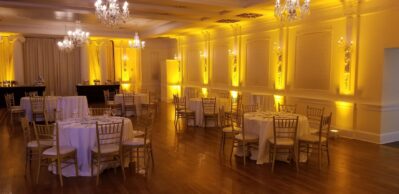Elegant banquet hall with round tables covered in white cloths, gold chairs, wooden floor, and warm yellow uplighting. Chandeliers hang from the ceiling, creating a formal atmosphere—perfect for events with dj services Suffolk and Nassau County, NY.