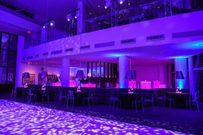 A modern NY,NY event space lit with vibrant purple and blue lighting, featuring tables with candles, transparent chairs, a sleek bar area, high ceilings, and a mezzanine—ideal for entertainment services Suffolk and Nassau County.