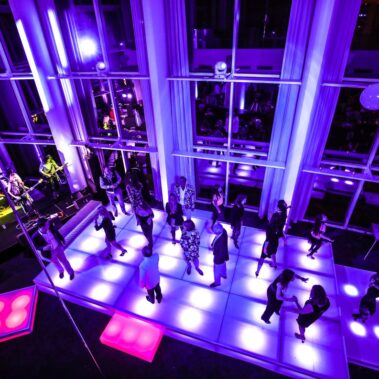 A group of people dance on a brightly lit, color-changing floor in a modern venue with vibrant purple lighting. A live band performs on stage to the left, showcasing top entertainment services Suffolk and Nassau County offer.