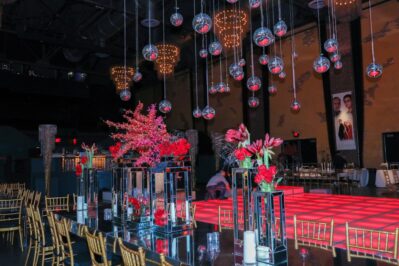 Decorated event space in NY with gold chairs, mirrored tables, and vibrant red floral centerpieces. Glass orbs hang above a glowing red checkered dance floor. Exposed brick walls and framed photos complete this stunning entertainment services Suffolk and Nassau County, NY setting.
