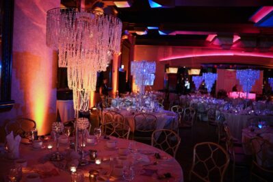 Elegant banquet hall with round tables set for an event, featuring tall crystal centerpieces, candlelight, and dramatic colored lighting in purple, orange, and blue hues. Perfect for entertainment services Suffolk and Nassau County, NY, with a luxurious festive atmosphere.