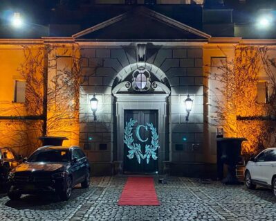 A grand building entrance at night in NY, illuminated by exterior lights. Two cars flank a red carpet leading to double doors with a wreath and the letter "C." Perfect for dj services Suffolk and Nassau County, NY. Cobblestone driveway in foreground.