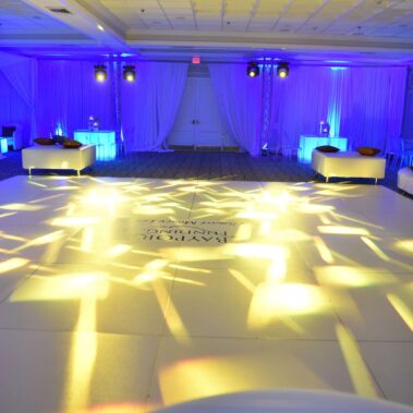 A large, elegant ballroom in NY with white sofas and cocktail tables, illuminated by blue and yellow stage lights that create patterns on a spacious dance floor. Draped white curtains line the walls, reflecting upscale entertainment services in Suffolk and Nassau County.