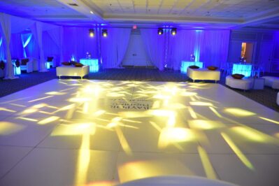 A large, elegant ballroom in NY with white sofas and cocktail tables, illuminated by blue and yellow stage lights that create patterns on a spacious dance floor. Draped white curtains line the walls, reflecting upscale entertainment services in Suffolk and Nassau County.