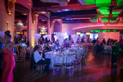A formal event with elegantly dressed guests seated at round tables in a dimly lit, ornately decorated ballroom featuring chandeliers, colorful lighting, and people mingling—enhanced by top DJ services Suffolk and Nassau County.