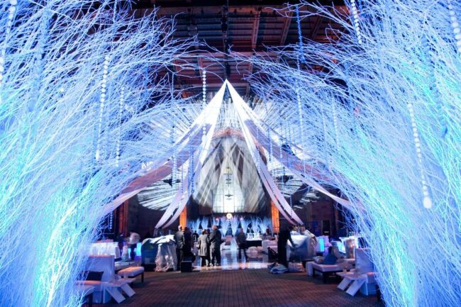 An indoor event space in NY,NY features glowing white branches and draped white fabric, creating a winter wonderland atmosphere. Guests gather under dramatic blue lighting—perfect for entertainment services in Suffolk and Nassau County.