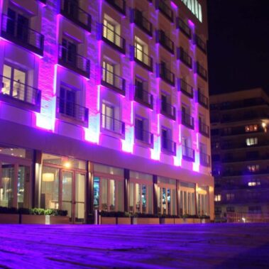 A modern multi-story building is illuminated with vibrant purple lights at night. Two people stand on the right, near the wooden pathway leading past the hotel—perfect for events featuring top DJ services in Suffolk and Nassau County, NY. Other buildings are visible in the background.