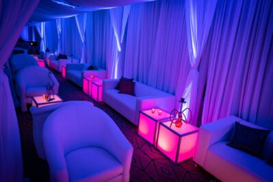 A lounge area with white sofas, glowing pink cube tables, sheer curtains, and dim blue-purple lighting creates an upscale vibe. Hookahs and candles adorn the tables—perfect for events with DJ services in Suffolk and Nassau County, NY.