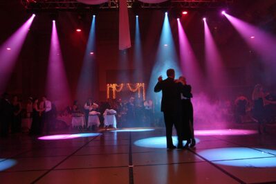 A couple dances in the center of a dimly lit ballroom under colorful spotlights, while people sit at round tables and watch—an unforgettable moment made possible by DJ services Suffolk and Nassau County, NY.