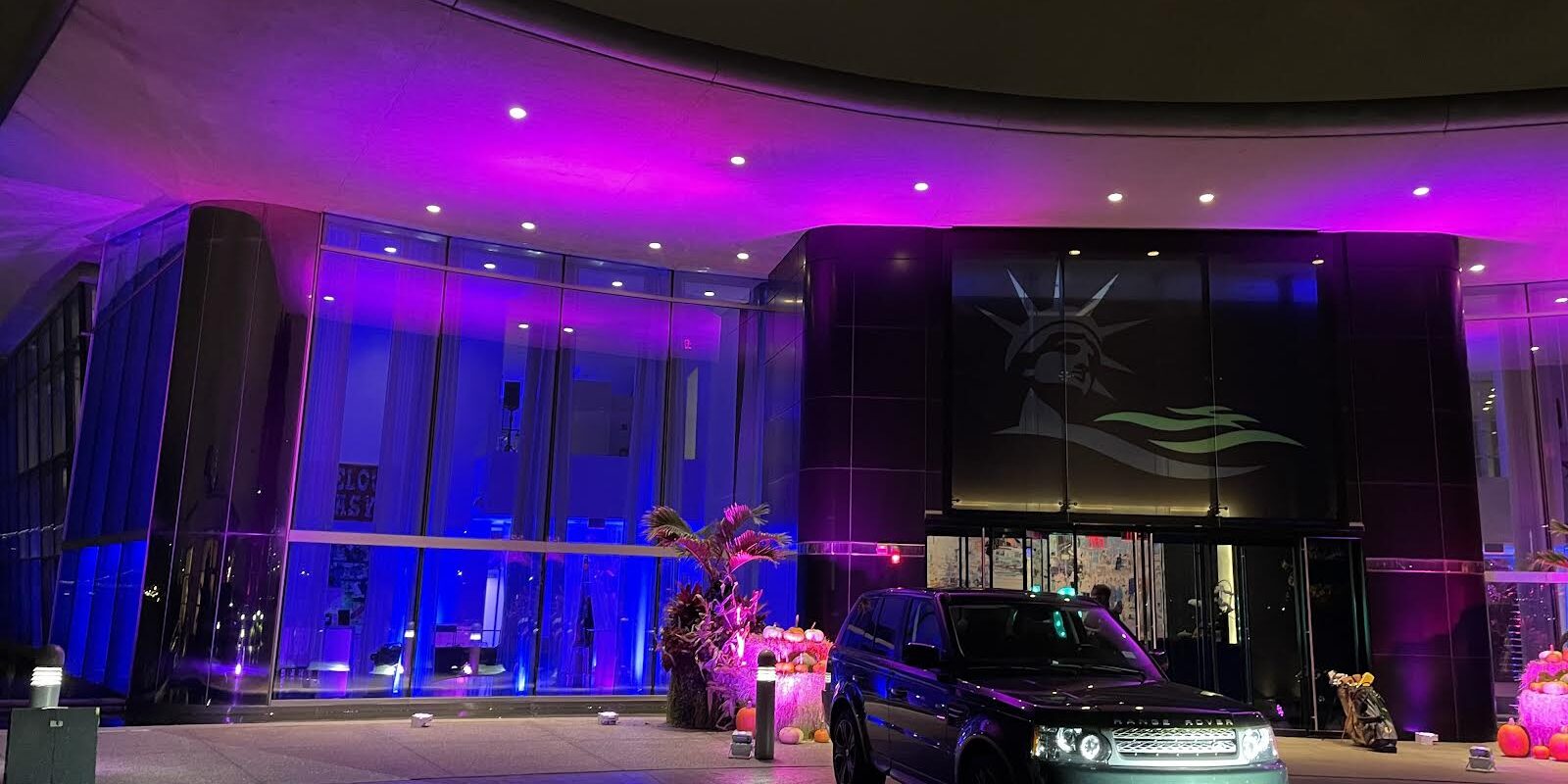 A modern building with a glowing pink and purple exterior is shown at night. Above the entrance, a large image of the Statue of Liberty’s face hints at top entertainment services in Suffolk and Nassau County, NY, with cars parked out front and orange decorations by the doors.