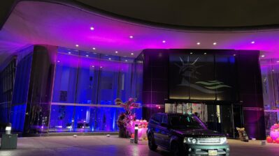 A modern building with a glowing pink and purple exterior is shown at night. Above the entrance, a large image of the Statue of Liberty’s face hints at top entertainment services in Suffolk and Nassau County, NY, with cars parked out front and orange decorations by the doors.