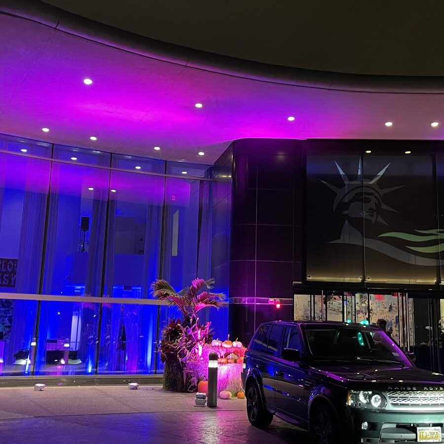 A modern building with a glowing pink and purple exterior is shown at night. Above the entrance, a large image of the Statue of Liberty’s face hints at top entertainment services in Suffolk and Nassau County, NY, with cars parked out front and orange decorations by the doors.