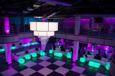A modern NY event space with a large checkered dance floor, white seating, glowing green and purple accent lighting, round tables, curved couches, and a central chandelier—perfect for parties with DJ services in Suffolk and Nassau County, NY.