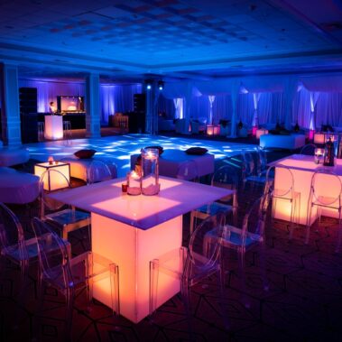 A modern NY,NY event space with glowing tables and chairs, illuminated in blue and purple. A lit dance floor and DJ booth set the scene for a stylish party, perfect for entertainment services in Suffolk and Nassau County.