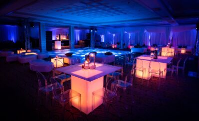 A modern NY,NY event space with glowing tables and chairs, illuminated in blue and purple. A lit dance floor and DJ booth set the scene for a stylish party, perfect for entertainment services in Suffolk and Nassau County.