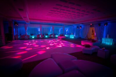 A spacious event room with white lounge furniture, draped curtains, and a large dance floor, illuminated by vibrant pink and blue lights—perfect for entertainment services in Suffolk and Nassau County.