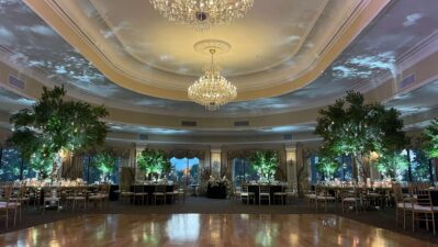 Elegant ballroom with large chandeliers, a polished dance floor, round tables set for an event, tall greenery decorations, and soft lighting—perfect for NY celebrations featuring top entertainment services Suffolk and Nassau County has to offer.