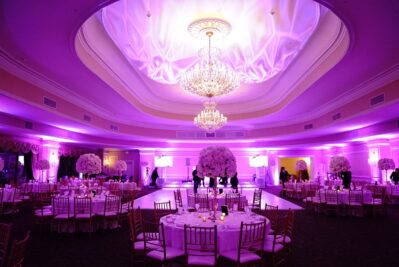 Elegant banquet hall decorated for an event with purple lighting, round tables with white linens and tall floral centerpieces, gold chairs, and large crystal chandeliers—perfect for entertainment services Suffolk and Nassau County.