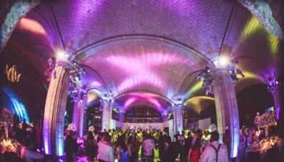 A large, elegant hall with high vaulted ceilings, colorful purple and yellow lighting, and a formally dressed crowd—possibly at a wedding or celebration in NY, featuring top entertainment services Suffolk and Nassau County. Tables with flowers line the space.