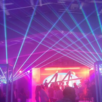 People dance in a club or event space in NY as blue laser lights crisscross above. A large, illuminated screen displays a colorful graphic, while top dj services Suffolk and Nassau County, NY provide vibrant beats for the crowd.