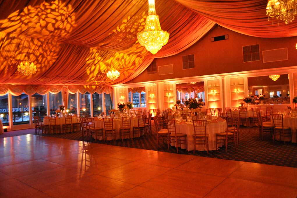 A large, elegant banquet hall in NY with round tables set for an event, golden chandeliers, and warm orange lighting reflecting off draped ceilings—perfect for entertainment services in Suffolk and Nassau County, creating a festive and inviting atmosphere.