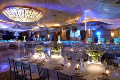 Elegant banquet hall in NY decorated for a formal event, featuring draped white fabric on the ceiling, a large leaf-shaped chandelier, glowing centerpieces with white flowers, and tables set with fine dinnerware and glassware. DJ services Suffolk and Nassau County available.