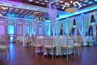 Elegant banquet hall with round tables covered in white tablecloths, gold chairs, tall crystal centerpieces, wooden floor, large windows with drapes, and blue ambient lighting—perfect for events featuring top entertainment services Suffolk and Nassau County.