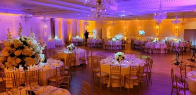 Elegant banquet hall in NY decorated for an event with round tables, gold chairs, white floral centerpieces, and warm yellow lighting. Crystal chandeliers hang from the ceiling, with a stage area ready for entertainment services Suffolk and Nassau County, NY.