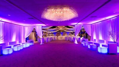 Elegant banquet hall with purple lighting, white draped curtains, a crystal chandelier, modern lounge seating, and festively set tables—perfect for upscale events with premium entertainment services in Suffolk and Nassau County, NY.