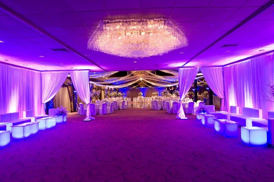 Elegant banquet hall with purple lighting, white draped curtains, a crystal chandelier, modern lounge seating, and festively set tables—perfect for upscale events with premium entertainment services in Suffolk and Nassau County, NY.