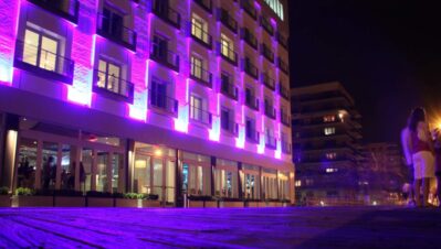 A modern multi-story building in NY is illuminated with vibrant purple lights at night. People stand on the sidewalk nearby, enjoying entertainment services Suffolk and Nassau County, while other buildings are visible in the background.