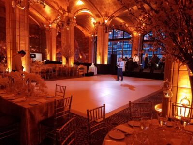 Elegant ballroom with golden lighting, decorated tables, and an empty dance floor. A small group gathers as a band sets up, while a woman stands alone beneath tall arched windows. Perfect for events featuring top DJ services in Suffolk and Nassau County, NY.