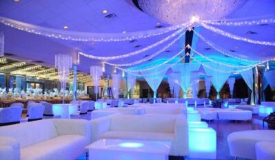 Elegant event hall with white sofas, glowing blue tables, hanging string lights, and crystal decorations, ideal for luxurious celebrations. Enhance your experience with entertainment services Suffolk and Nassau County under a grand chandelier.