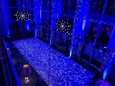 A modern event space is illuminated with blue lighting and patterned light spots on the floor, ideal for entertainment services Suffolk and Nassau County, NY. Tall windows and decorative lamps create a perfect setting for any occasion or dj services Suffolk and Nassau County.