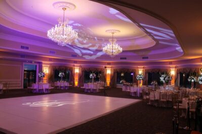 Elegant ballroom with chandeliers, purple and gold lighting, round tables with flower centerpieces, and a large white dance floor in the center—perfect for a formal event or wedding reception featuring top DJ services Suffolk and Nassau County.