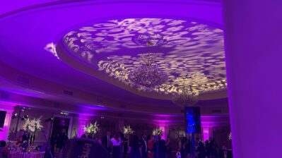 A banquet hall with ornate chandeliers and decorative purple lighting patterns projected onto the ceiling. Guests are mingling near elegant floral centerpieces on round tables, enjoying top-tier entertainment services Suffolk and Nassau County, NY.